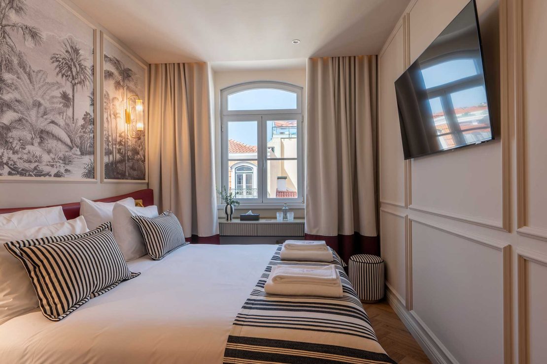 Hotel bedroom with white bedding and black‑and‑white striped pillows, palm mural artwork, and a window letting in natural light beside a wall-mounted TV.
