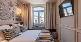 Hotel bedroom with white bedding and black‑and‑white striped pillows, palm mural artwork, and a window letting in natural light beside a wall-mounted TV.