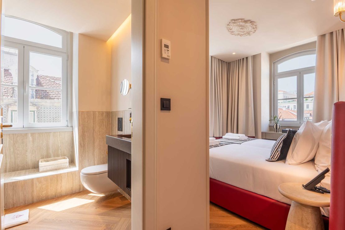 Split view of a hotel room and bathroom: right side shows a bed with white linens, left side shows a marble-tiled bathroom with a toilet and window.
