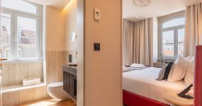 Split view of a hotel room and bathroom: right side shows a bed with white linens, left side shows a marble-tiled bathroom with a toilet and window.
