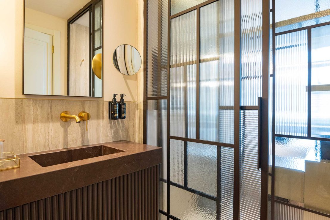 Contemporary bathroom vanity with dark brown marble counter, gold faucet, round mirror, and textured glass partition.