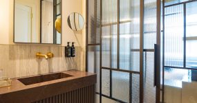 Contemporary bathroom vanity with dark brown marble counter, gold faucet, round mirror, and textured glass partition.