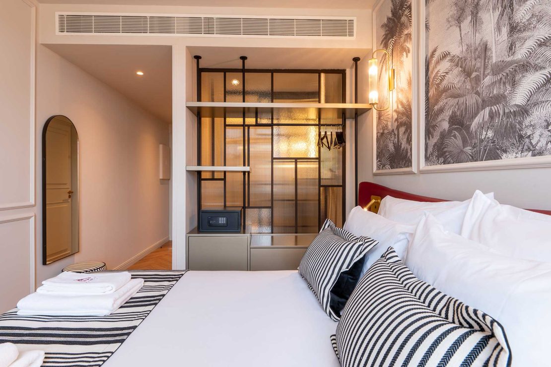 Modern hotel bedroom with white bedding, striped cushions, and a glass-panel closet area against a palm-patterned wall.