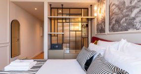 Modern hotel bedroom with white bedding, striped cushions, and a glass-panel closet area against a palm-patterned wall.