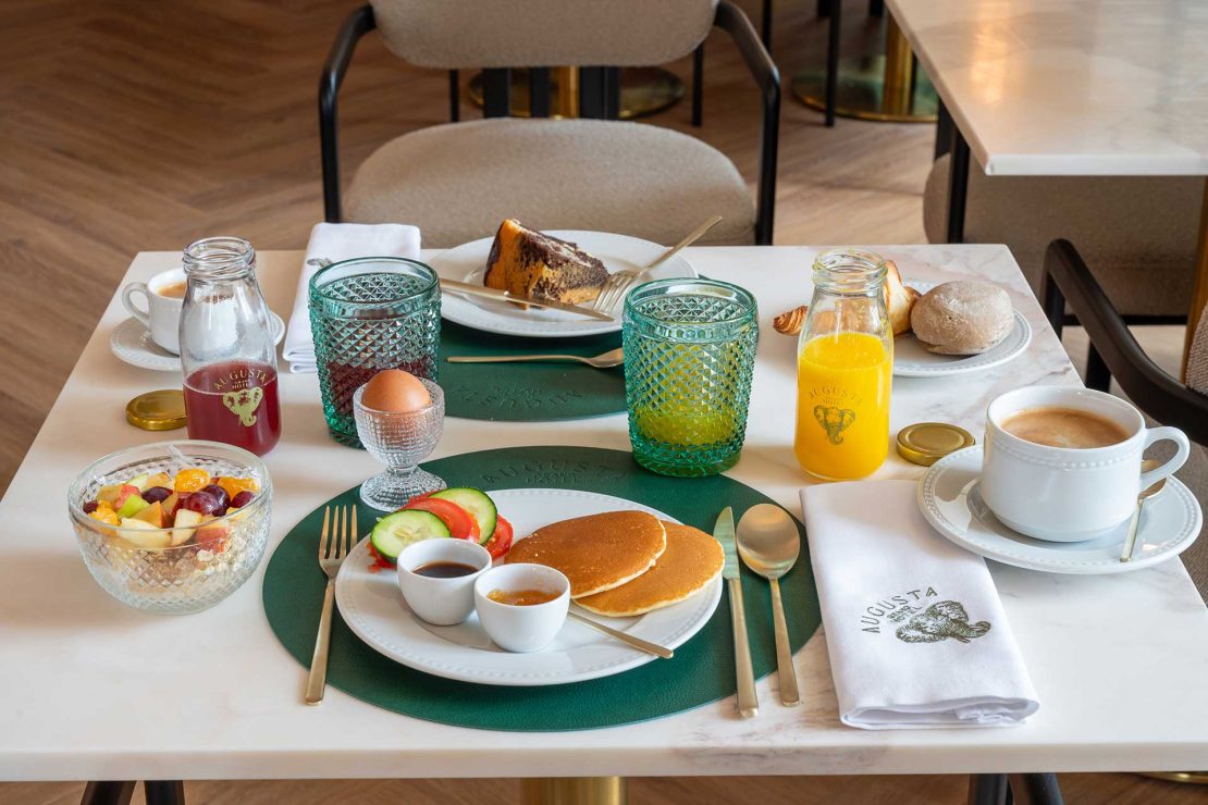 Table set for breakfast with pancakes, fruit salad, juice, coffee, bread rolls, croissant and a green placemat centerpiece on a marble table.