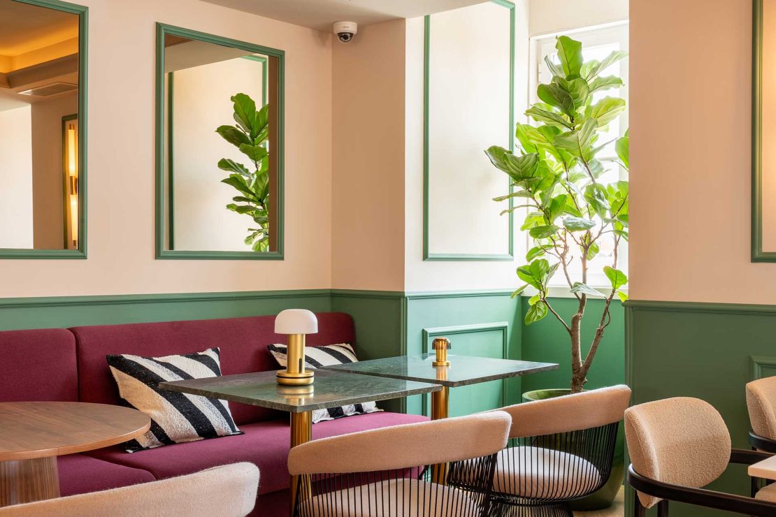 Cozy lounge area with a pink sofa, striped cushions, and round wooden tables, set against green wainscoted walls and tall leafy plant.
