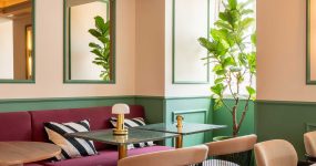 Cozy lounge area with a pink sofa, striped cushions, and round wooden tables, set against green wainscoted walls and tall leafy plant.
