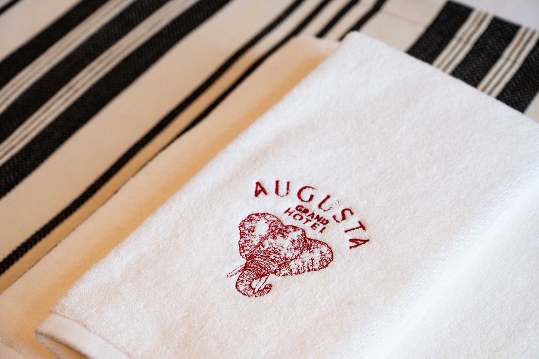 White towel with a red Augusta Grand Hotel logo and elephant illustration, on a beige-and-black striped surface.