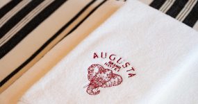 White towel with a red Augusta Grand Hotel logo and elephant illustration, on a beige-and-black striped surface.