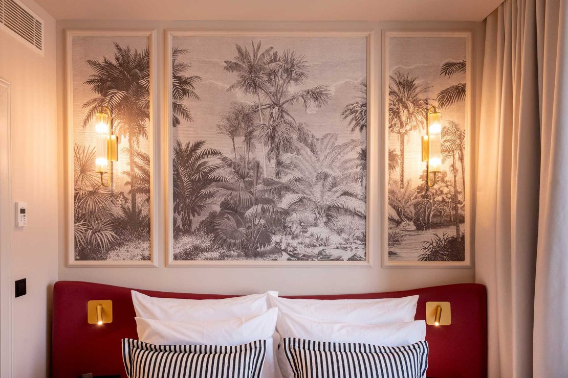 Triptych palm tree scene above a red headboard with white pillows and two warm wall lamps on either side. Curtains frame the right side of the scene.