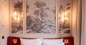 Triptych palm tree scene above a red headboard with white pillows and two warm wall lamps on either side. Curtains frame the right side of the scene.