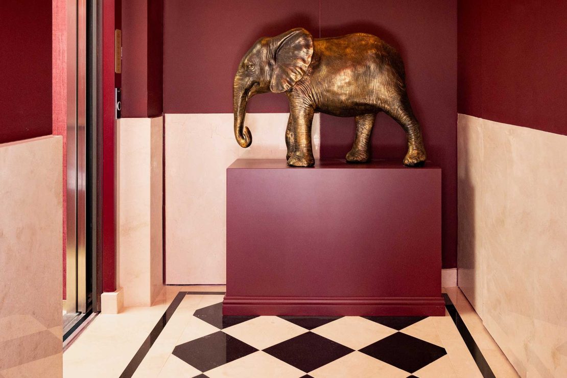 Bronze elephant statue on a burgundy pedestal in a red-walled foyer with a black-and-white checkered floor.
