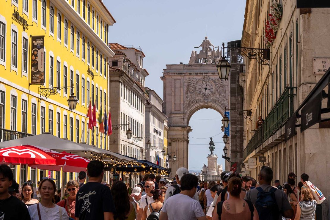 Bustling street scene with a bright yellow building on the left, outdoor cafés with red umbrellas, and a historic arch in the distance amid a crowd of people.