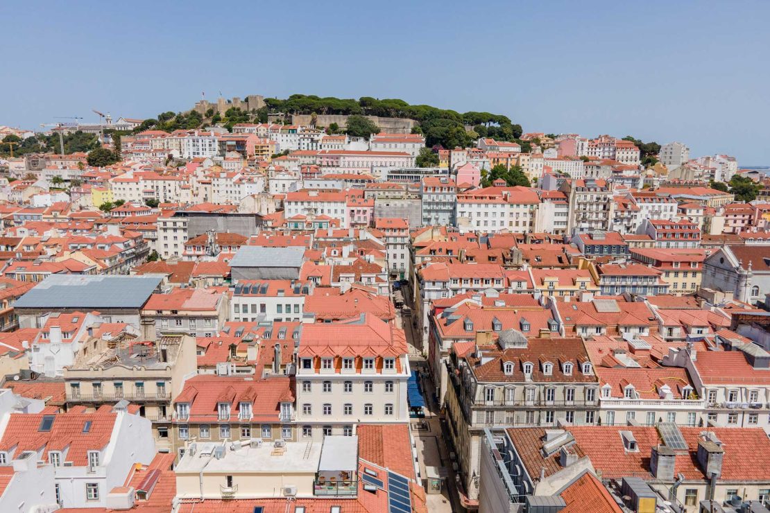 Aerial view of a dense town with red-tiled roofs and a hilltop fortress in the distance under a clear blue sky.