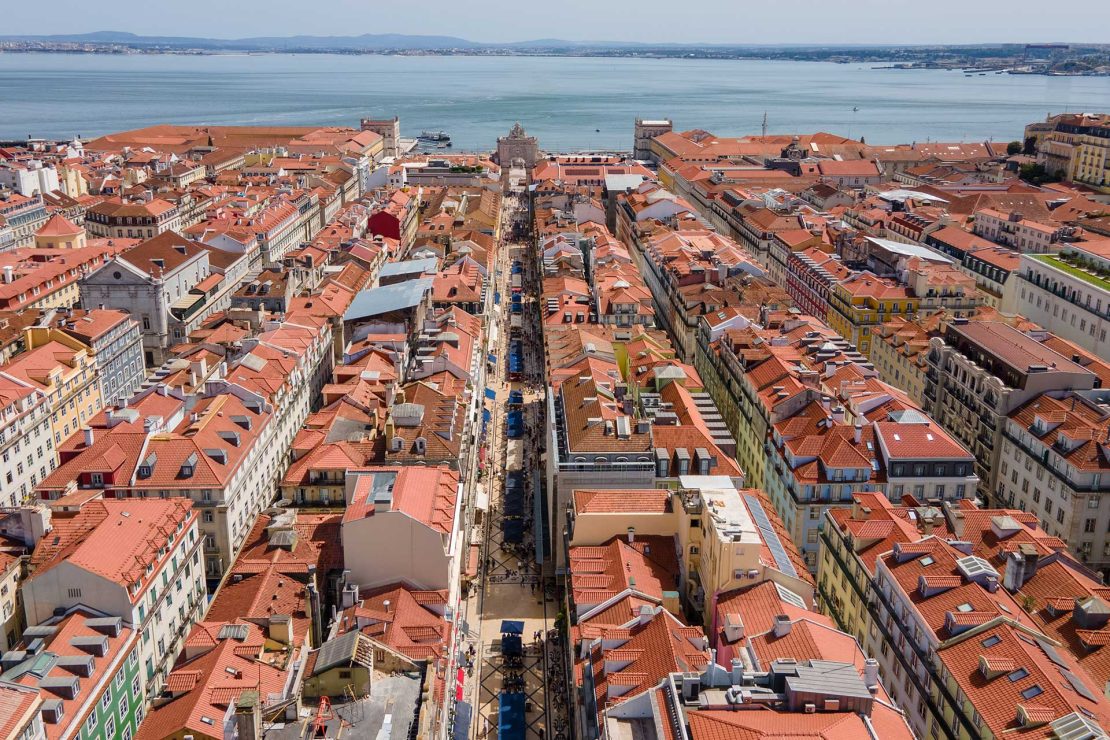 Aerial view of a dense city with red-tiled rooftops along a central street leading to the river.