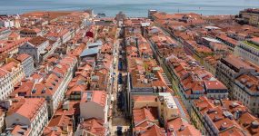 Aerial view of a dense city with red-tiled rooftops along a central street leading to the river.