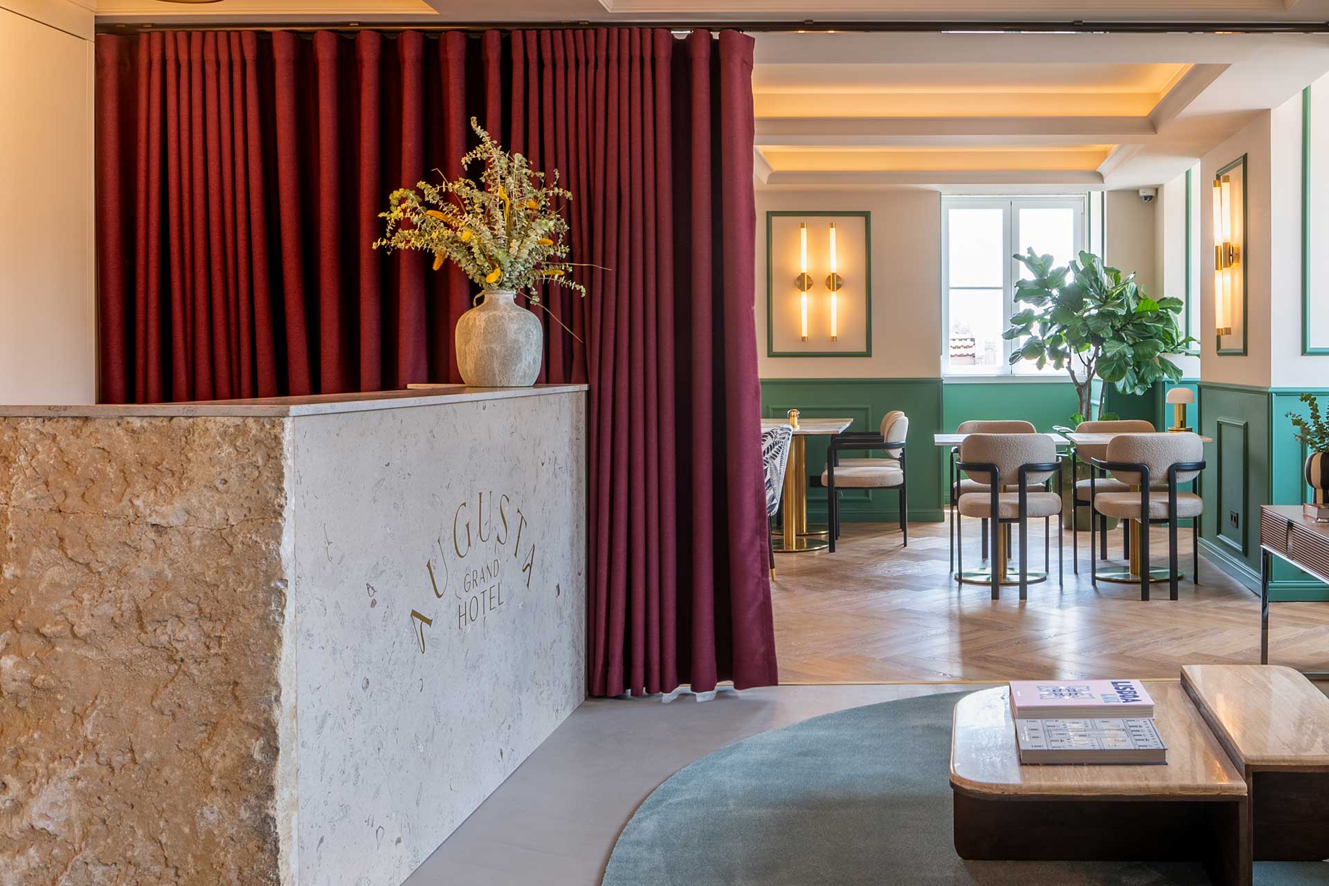 Hotel lobby reception with a marble counter, burgundy curtains, and a tall vase of dried flowers.