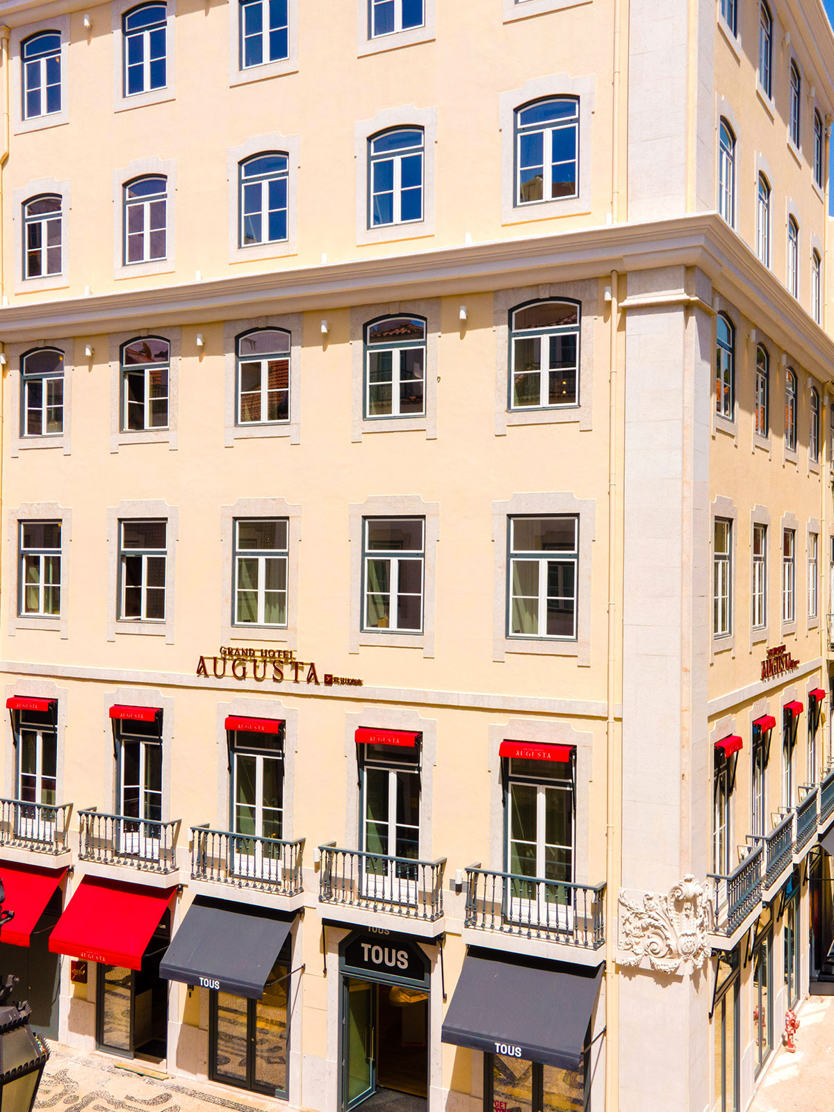 Facade of a light-yellow hotel building with white-framed arched windows and red_storefront awnings along the ground floor.