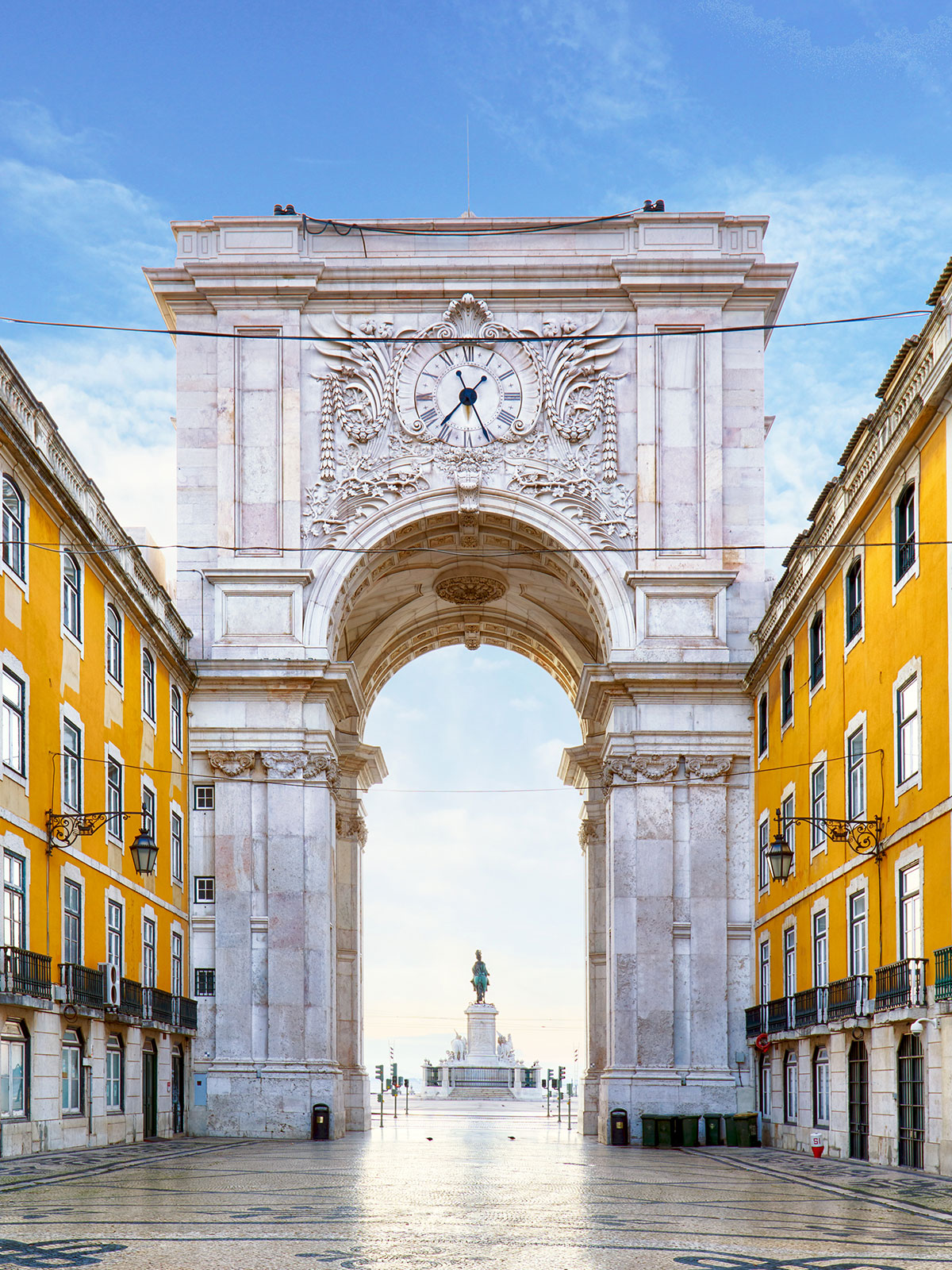 Ornate stone arch with a clock at the top, yellow buildings on both sides, and a statue in the square beyond.